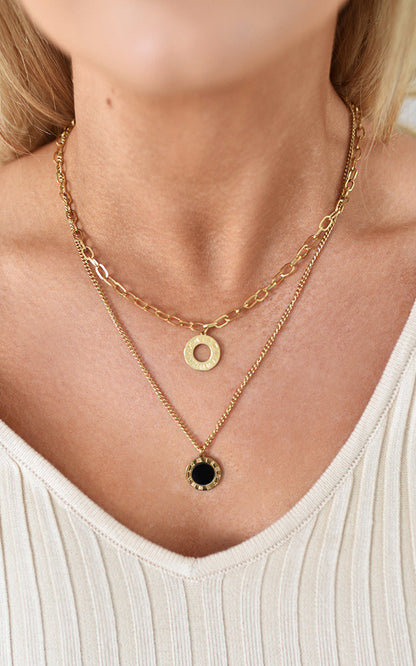 Two-layer gold necklace featuring a delicate chain with a black circular pendant and a shorter chain with a gold ring pendant. Stylish and elegant jewelry piece by Clarizzma.